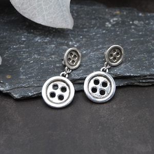 Puede incluir: Un par de pendientes de botón plateados. Cada pendiente presenta un pequeño botón y un dije de botón más grande que cuelga. Los botones tienen cuatro agujeros y están sobre un fondo oscuro.