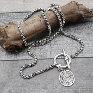 May include: A silver chain necklace with a toggle clasp and a round pendant with an intricate design.