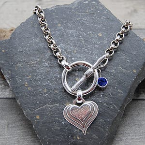 May include: A silver-toned necklace with a toggle clasp and a heart pendant. The heart pendant has a layered design with a dotted pattern. A small blue gemstone charm hangs near the clasp. The necklace is displayed on a dark gray stone.