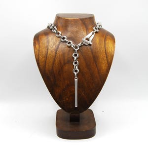 Puede incluir: Un collar de cadena de plata con un colgante largo y delgado que cuelga de él. El collar está en un expositor de madera.