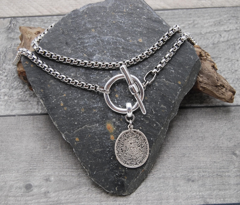 May include: A silver chain necklace with a toggle clasp and a round pendant with an intricate design. The necklace is layered on a gray stone surface.