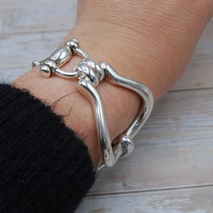 May include: A silver-toned bracelet with a unique design. The bracelet features a series of curved, connected metal pieces, creating an open, geometric shape. The metal has a polished, reflective surface. The bracelet is worn on a wrist.