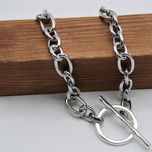 May include: A silver chain necklace with a large, round toggle clasp. The chain is made of thick, oval-shaped links.