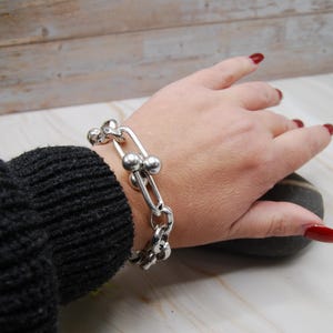 Könnte beinhalten: Ein silberfarbenes Armband mit einem Kettenglied-Design und kugelförmigen Akzenten. Das Armband wird am Handgelenk getragen, wobei ein dunkelgrauer Pulloverärmel sichtbar ist. Der Hintergrund ist ein helles Holzmaserungsmuster.