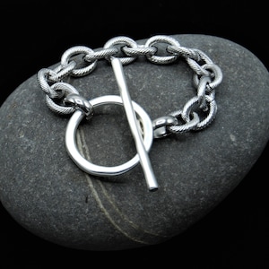 Puede incluir: Un brazalete de cadena de plata con cierre de palanca. El brazalete está hecho de una cadena gruesa y texturizada y tiene un diseño simple y moderno.