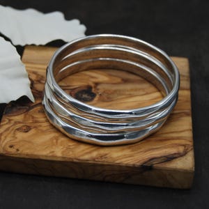 May include: Four silver bangles with a textured, wave-like design, displayed on a small wooden board. The bangles are stacked together. The board has a natural wood grain. The background is dark.