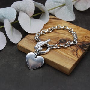 Op de afbeelding: Een zilverkleurige armband met een hartvormige hanger en een tuimelsluiting. De armband ligt op een klein, vierkant houten blok met een natuurlijke houtnerf. Eucalyptustakken zijn op de achtergrond.