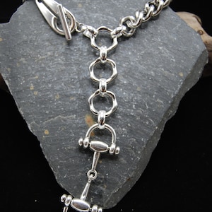 May include: A silver-toned bracelet with a toggle clasp and a unique equestrian-themed design. The bracelet features a chain link, connected to a series of rings and a horse bit pendant. The jewelry is displayed on a gray stone surface.