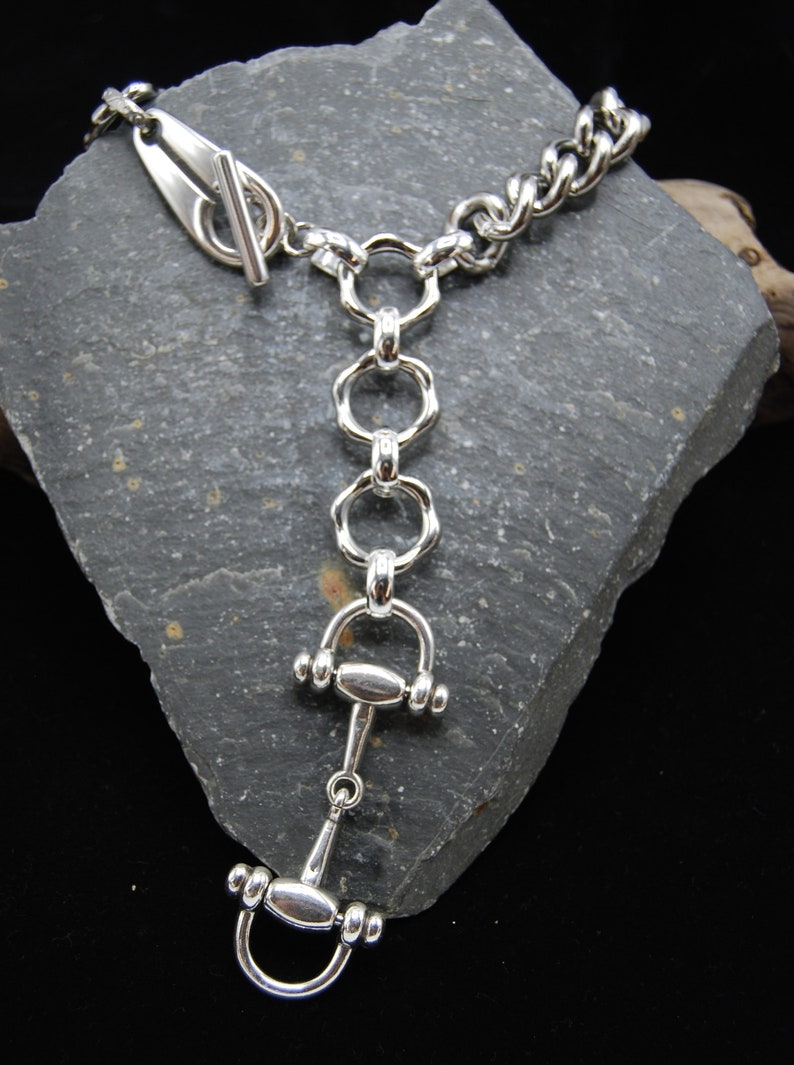 May include: A silver-toned bracelet with a toggle clasp and a unique equestrian-themed design. The bracelet features a chain link, connected to a series of rings and a horse bit pendant. The jewelry is displayed on a gray stone surface.