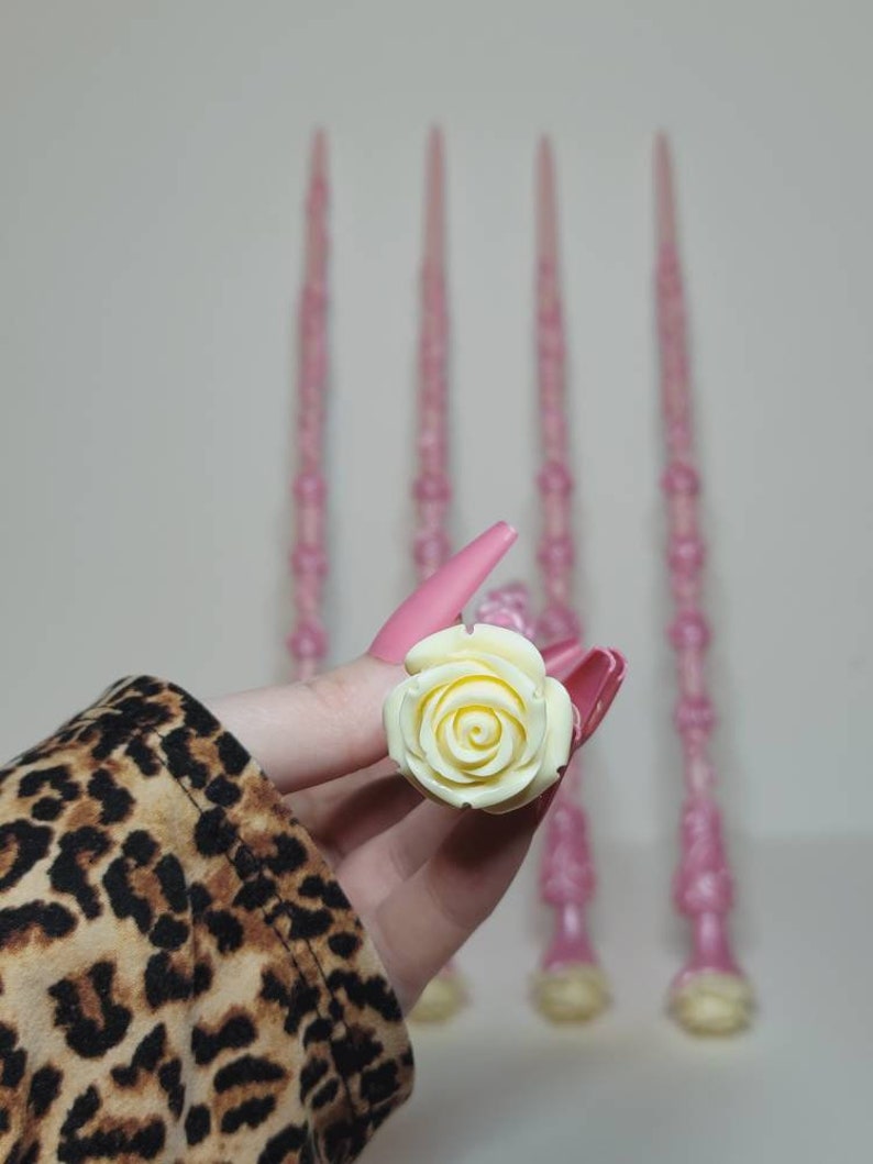 May include: Four pink wands with white rose-shaped tops. The wands are made of a sparkly material and have a delicate, feminine look.