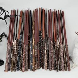May include: A collection of 25 wooden wands with various designs and colors, including brown, black, gold, and silver. The wands are arranged in a row on a white surface.
