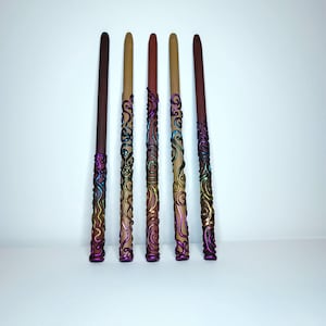May include: Six handcrafted wands with a fantasy theme. The wands have a dark brown handle and a light brown shaft. The handles are decorated with iridescent swirls of purple, blue, and gold. The wands are arranged vertically against a white background.