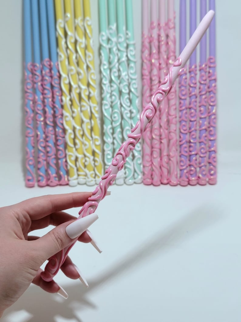May include: A pink wand with a decorative, swirling pattern, held in front of a collection of colourful wands. The wand has a detailed, raised design in a darker pink. Other wands are in shades of blue, yellow, green, pink, and purple.