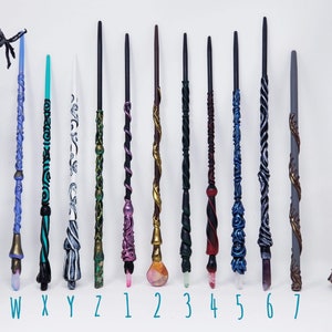 May include: A selection of handcrafted wands in diverse colours and designs. Each wand has unique details, including carved patterns, crystal accents, and metallic finishes. The wands are labelled with letters and numbers.