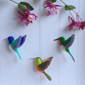 May include: Three felt hummingbird ornaments in different colors. The hummingbirds are green, blue, and orange with brown accents. They are hanging on a white wall.