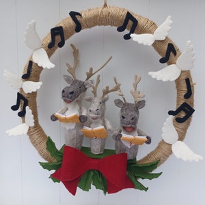 Op de afbeelding: Een kerstkrans van vilt met drie rendieren die kerstliederen zingen. De krans is versierd met witte viltvleugels en zwarte muzieknoten. De rendieren houden bladmuziek vast en dragen rode strikken.