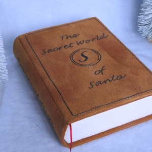 May include: A brown book with a gold-trimmed spine and the title "The Secret World of Santa" in black lettering. The book has a white page edge and a red ribbon bookmark.