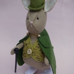 May include: A plush toy rabbit wearing a green top hat, a green cape, and a yellow and red patterned vest. The rabbit is holding a black walking stick.