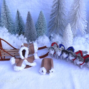 Puede incluir: Un trineo de fieltro marrón con una capa con capucha de fieltro marrón y un equipo de ocho perros husky de fieltro tirando del trineo. Los perros llevan arneses rojos y azules. La escena está ambientada en un país de las maravillas invernal nevado con pinos blancos escarchados.