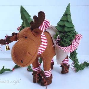 Pode incluir: Um alce de feltro marrom carregando uma pequena árvore de Natal de feltro verde nas costas. O alce está usando um cachecol listrado vermelho e branco e tem um sino pendurado no pescoço. O alce está em pé em uma superfície branca com uma guirlanda verde arrastando-se atrás dele.