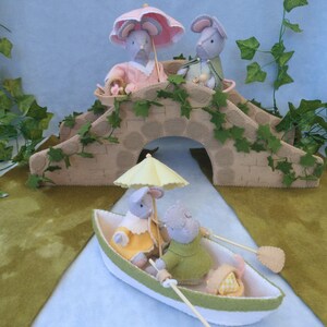 In this picture: a felt handmade  bridge, old style, with ivy vines along the upper side. The bridge is decorated with a stone pattern and is standing on two green waterfronts above blue water. This bridge is part of a Summer sewing project with elegant mice dressed in vintage clothing, Father, mother and two children in a boat.  All made of wool felt with a PDF hand sewing pattern from WoolFeltwonders.