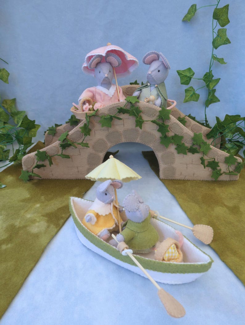 In this picture: a felt handmade  bridge, old style, with ivy vines along the upper side. The bridge is decorated with a stone pattern and is standing on two green waterfronts above blue water. This bridge is part of a Summer sewing project with elegant mice dressed in vintage clothing, Father, mother and two children in a boat.  All made of wool felt with a PDF hand sewing pattern from WoolFeltwonders.