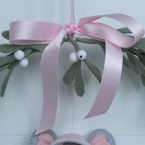 In this picture: a details of asweet handmade felt mouse with pink dress, white cape and pink and white bonnet, holding a bunch of felt mistletoe with white berries between her hands. She is hanging in a wreath with pink bow and felt mistletoe with white berries, on a white surface. This sweet grey mouse is made with a PDF hand sewing pattern from WoolFeltWonders by Tintangel.