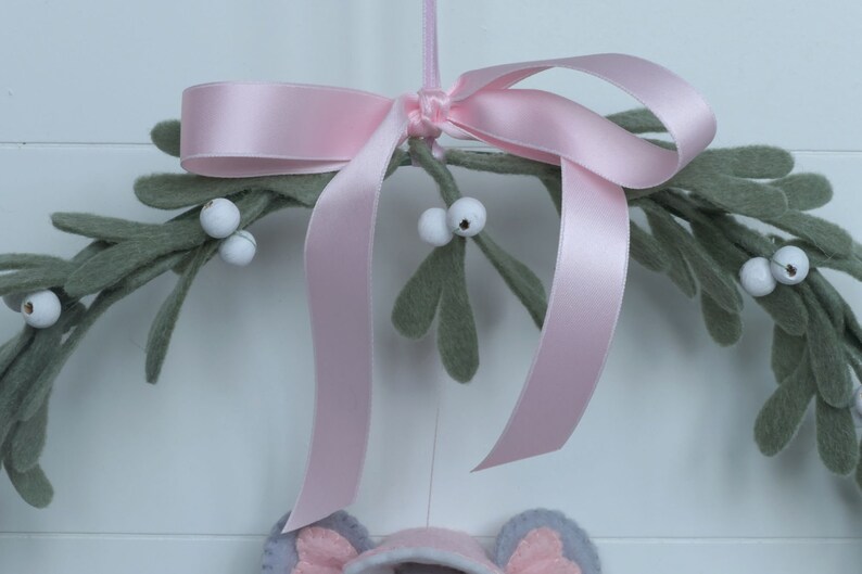In this picture: a details of asweet handmade felt mouse with pink dress, white cape and pink and white bonnet, holding a bunch of felt mistletoe with white berries between her hands. She is hanging in a wreath with pink bow and felt mistletoe with white berries, on a white surface. This sweet grey mouse is made with a PDF hand sewing pattern from WoolFeltWonders by Tintangel.