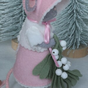 In this picture: a sweet handmade felt mouse with pink dress, white cape and pink and white bonnet, holding a bunch of felt mistletoe with white berries between her hands. She is standing on front of two fake trees on a white surface. This sweet grey mouse is made with a PDF hand sewing pattern from WoolFeltWonders by Tintangel.