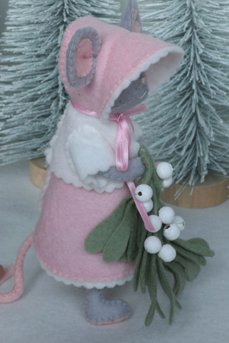 In this picture: a sweet handmade felt mouse with pink dress, white cape and pink and white bonnet, holding a bunch of felt mistletoe with white berries between her hands. She is standing on front of two fake trees on a white surface. This sweet grey mouse is made with a PDF hand sewing pattern from WoolFeltWonders by Tintangel.