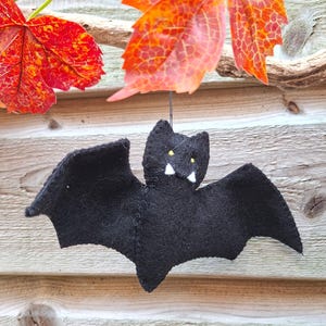 May include: A black felt bat ornament featuring small yellow embroidered eyes and two white felt fangs. The bat is suspended by a thin black string, showcasing a soft texture and pointed wing tips, designed as a hanging decoration for seasonal displays.