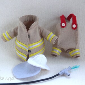 In this picture: a firefigther costumes for felt toy animals, made with a PDF handsewing pattern from WoolFeltWonders, part of a series with toy animal and clothing patterns to mix and match, to portray hobby or profession, for decoration and play. this set has a sand-colored jacket and trousers with white helmet.  All clothes have reflecting stripes in yellow and silver. A waterhose is lying in front of the clothes.
