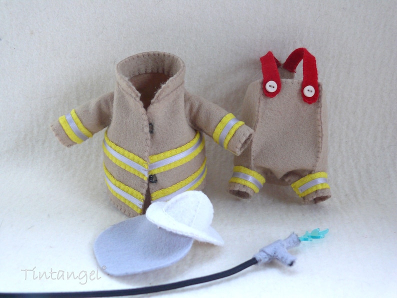In this picture: a firefigther costumes for felt toy animals, made with a PDF handsewing pattern from WoolFeltWonders, part of a series with toy animal and clothing patterns to mix and match, to portray hobby or profession, for decoration and play. this set has a sand-colored jacket and trousers with white helmet.  All clothes have reflecting stripes in yellow and silver. A waterhose is lying in front of the clothes.