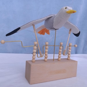 Op de afbeelding: Houten kinetische sculptuur van een zeemeeuw. De vogel heeft een grijs en wit vilten lichaam, zwarte vleugeltips en een oranje staart. Gemonteerd op een houten voet met houten kralen en messingdraad. De vleugels van de zeemeeuw zijn uitgespreid.