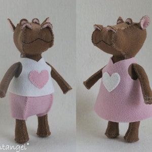 Op de afbeelding: Een bruin vilten nijlpaard dat een roze jurk draagt met een wit hart op de voorkant. Het nijlpaard heeft roze vilten oren en een wit vilten hart op de borst.
