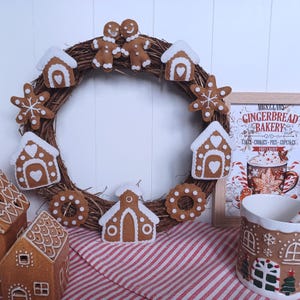 Op de afbeelding: Een bruine vilten gingerbreadkrans met witte vilten gingerbreadversieringen, waaronder huizen, sneeuwvlokken en een gingerbreadman en -vrouw. De krans hangt aan een witte muur. Een gingerbreadmok met een sneeuwvlokkenontwerp staat op de voorgrond.