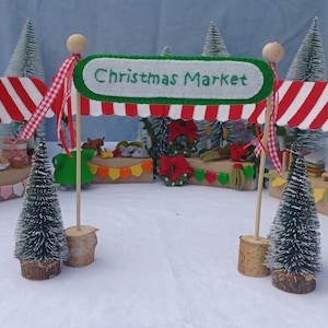 Op de afbeelding: Een miniatuur kerstmarktscène met rood-wit gestreepte luifels, kleine dennenbomen en viltversieringen. Het bord leest "Christmas Market". Twee muizen zijn gekleed in feestelijke kleding.