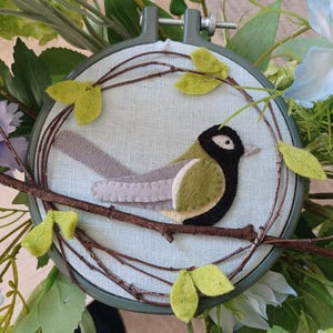 In this picture:  hand made wall decoration with a felt birds on linen in an embroidery hoop, the bird is a great  tit. The hoop is lying between flowers and leaves. This easy to make wall decoration is made with a PDF hand sewing pattern from WoolFeltWonders by Tintangel.
