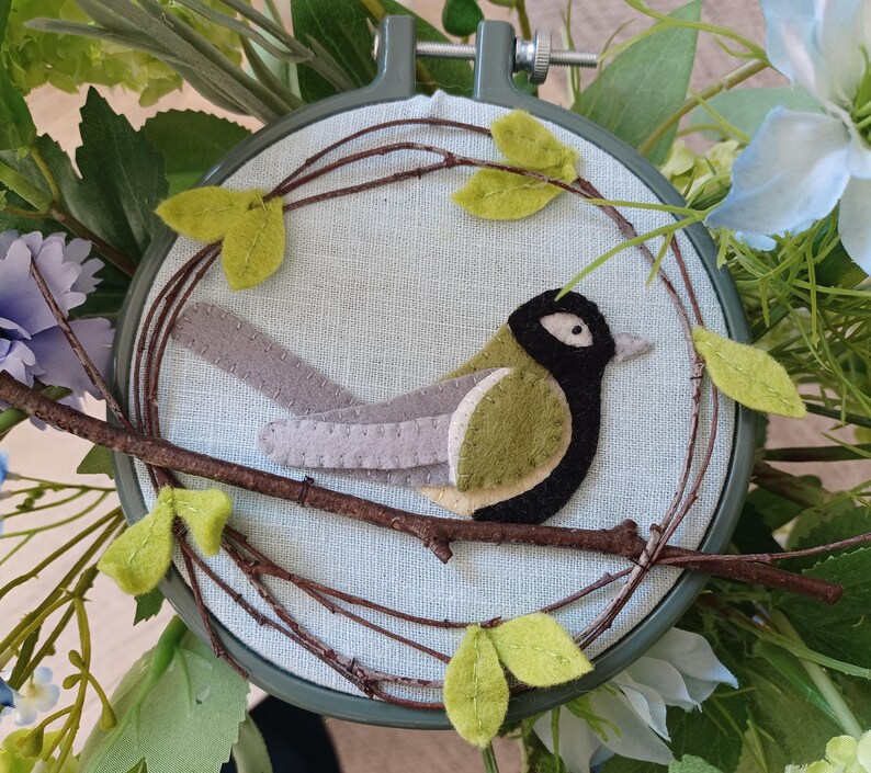 In this picture:  hand made wall decoration with a felt birds on linen in an embroidery hoop, the bird is a great  tit. The hoop is lying between flowers and leaves. This easy to make wall decoration is made with a PDF hand sewing pattern from WoolFeltWonders by Tintangel.
