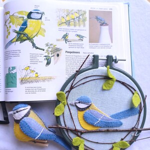 In this picture:  hand made wall decoration with a felt birds on linen in an embroidery hoop, the bird is a Bleut tit. The hoop is lying besides a book with a picture of the Bleu tit and a hand made felt Bleu tit in real life size on a white surface. This easy to make wall decoration is made with a PDF hand sewing pattern from WoolFeltWonders by Tintangel.