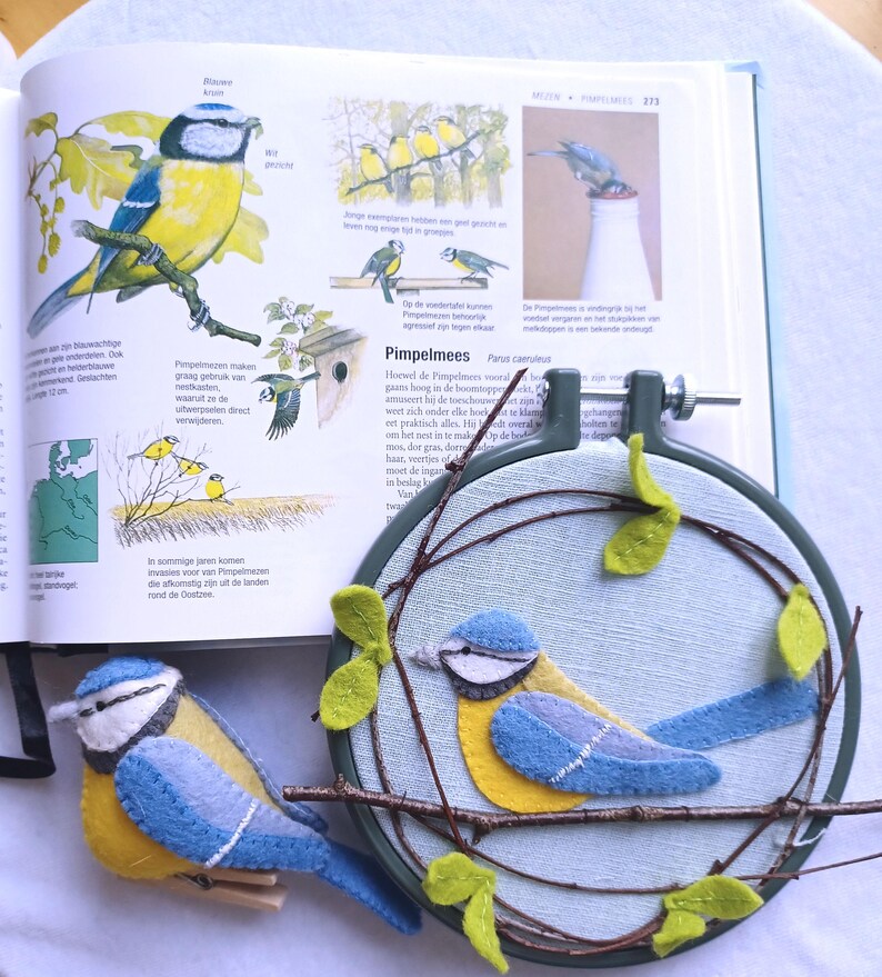 In this picture:  hand made wall decoration with a felt birds on linen in an embroidery hoop, the bird is a Bleut tit. The hoop is lying besides a book with a picture of the Bleu tit and a hand made felt Bleu tit in real life size on a white surface. This easy to make wall decoration is made with a PDF hand sewing pattern from WoolFeltWonders by Tintangel.