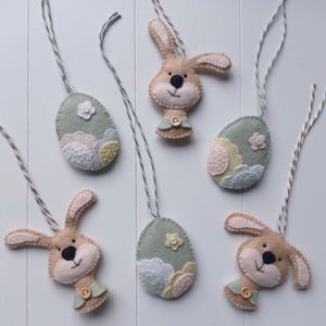 Op de afbeelding: Een verzameling handgemaakte paasornamenten. De ornamenten bevatten vilten konijntjes en eivormen in pastelkleuren, met bloemen- en wolkenachtige details. Elk ornament heeft een touwtje om op te hangen.
