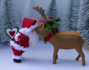 May include: A felt Santa Claus doll wearing a red suit with white trim and a white beard, holding a green heart with a red heart inside. The Santa doll is kissing a felt reindeer doll with brown fur, antlers, and a red and white checkered bow. The reindeer is wearing a red collar with a green holly sprig and a gold bell.