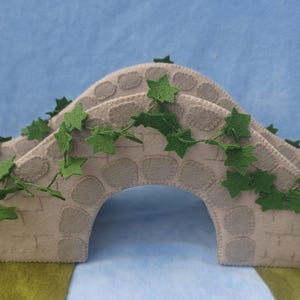 Könnte beinhalten: Eine beige Filzbrücke mit einem gebogenen Bogen, verziert mit grünem Efeu aus Filz. Die Brücke hat eine steinartige Textur und ist vor einem blau-grünen Hintergrund platziert, was eine skurrile, handgefertigte Ästhetik erzeugt.