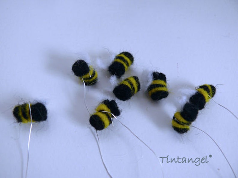 in this picture: 8 handmade felt bees, black with yellow stripes and white wings, all on a piece of thin silver wire. Thess bees are aprt of a PDF handsewing pattern from WoolFeltwonders to dress a felt toy animal as a bee keeper. the set contains, the clothing, bee hood, beehive, smoke pipe and these bees. Part of a series with felt toy animals and all kinds of clothing sets to mix and match to portray hobby or profession. Easy to make animals and clothing sets for decoration and play