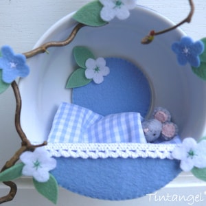 May include: A small, white mouse sleeps in a blue and white gingham bed inside a teacup. The teacup is decorated with blue felt flowers and leaves.