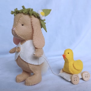 Puede incluir: Un conejito de fieltro marrón que lleva un chaleco blanco y una corona de hojas verdes tira de un pato de fieltro amarillo en un carro de madera con ruedas.