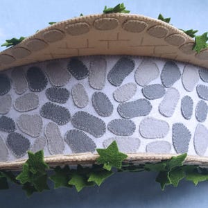 In this picture: a felt handmade  bridge, old style, with ivy vines along the upper side. The bridge is decorated with a stone pattern and is standing on two green waterfronts above blue water. This bridge is part of a Summer sewing project with elegant mice dressed in vintage clothing, Father, mother and two children in a boat.  All made of wool felt with a PDF hand sewing pattern from WoolFeltwonders.