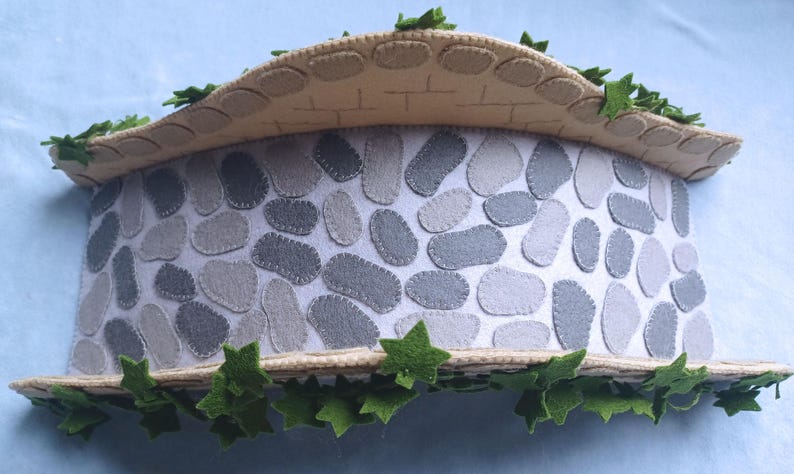 In this picture: a felt handmade  bridge, old style, with ivy vines along the upper side. The bridge is decorated with a stone pattern and is standing on two green waterfronts above blue water. This bridge is part of a Summer sewing project with elegant mice dressed in vintage clothing, Father, mother and two children in a boat.  All made of wool felt with a PDF hand sewing pattern from WoolFeltwonders.
