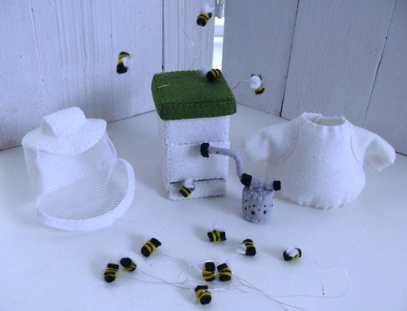 In this picture: a handmade set with clothing and accessoiries to dress a felt toy animal as a Beekeeper, with white jacket, bee hood, beehive, smoke pipe and many bees. All this is is made with PDF hand sewing pattern from WoolFeltWonders by Tintangel and is part of a series with felt toy animals and all kinds of clothing sets to mix and match to portray hobby or profession. Easy to make animals and clothing sets for decoration and play.